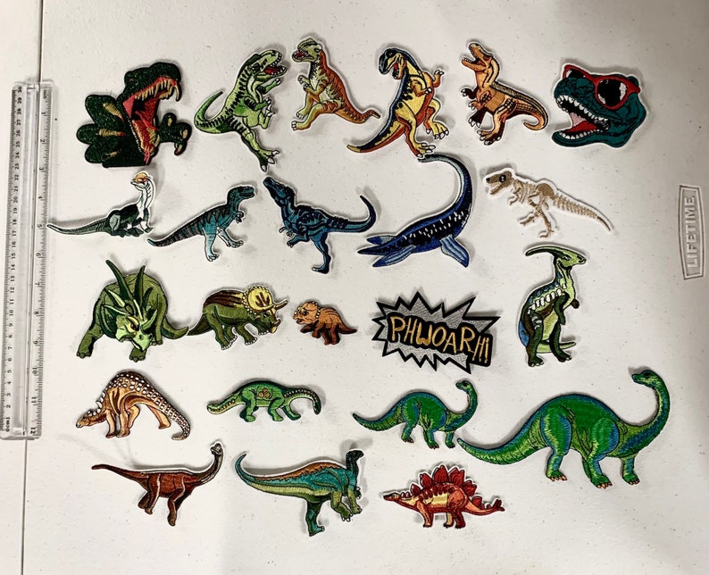 May include: A collection of 18 colorful dinosaur iron-on patches. The patches feature various dinosaurs in different poses, including a T-Rex, a Stegosaurus, and a Triceratops. The patches are perfect for adding a fun and unique touch to clothing, bags, and other accessories.