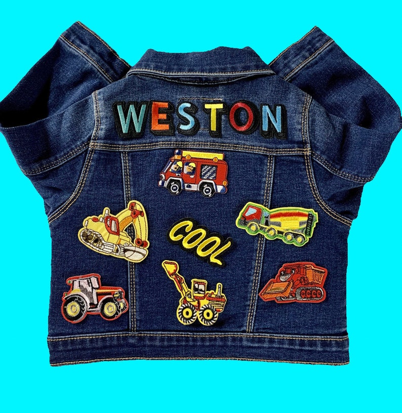 May include: A blue denim jacket with colorful patches of construction vehicles and the name "WESTON" on the back. The patches include a fire truck, a cement mixer, a backhoe, a bulldozer, and a tractor.