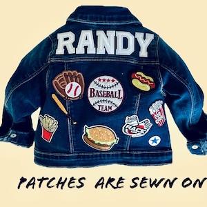 May include: A dark blue denim jacket with the name "RANDY" in white block letters across the back. The jacket is decorated with various patches, including a baseball, hot dog, and fries. The text "PATCHES ARE SEWN ON" is at the bottom.