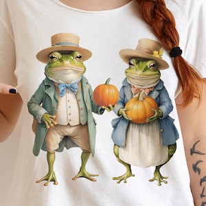 Puede incluir: Camiseta blanca con una ilustración caprichosa de dos ranas antropomórficas. Una rana lleva una pajarita azul y sostiene una calabaza, la otra lleva un abrigo azul y sostiene una calabaza. Ambas ranas llevan sombreros.