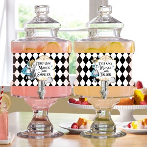 Puede incluir: Dos dispensadores de bebidas de vidrio con boquillas, decorados con patrones de cuadros negros y blancos y etiquetas con el tema de Alicia en el País de las Maravillas. Las etiquetas dicen "Este te hace más pequeño" y "Este te hace más alto".