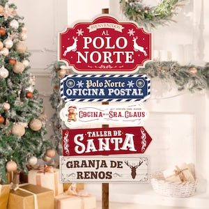 May include: A set of five wooden signs with red, blue, and white paint, decorated with snowflakes and reindeer. The signs point in different directions and feature the text "Bienvenidos al Polo Norte", "Polo Norte Oficina Postal", "Cocina de la Sra. Claus", "Taller de Santa", and "Granja de Renos".