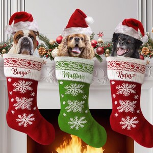 Puede incluir: Tres calcetines de Navidad rojos y verdes con copos de nieve blancos y ribete de piel. Cada calcetín tiene la cabeza de un perro asomando por la parte superior y un nombre bordado en la parte delantera: Bingo, Mullin y Rufus.