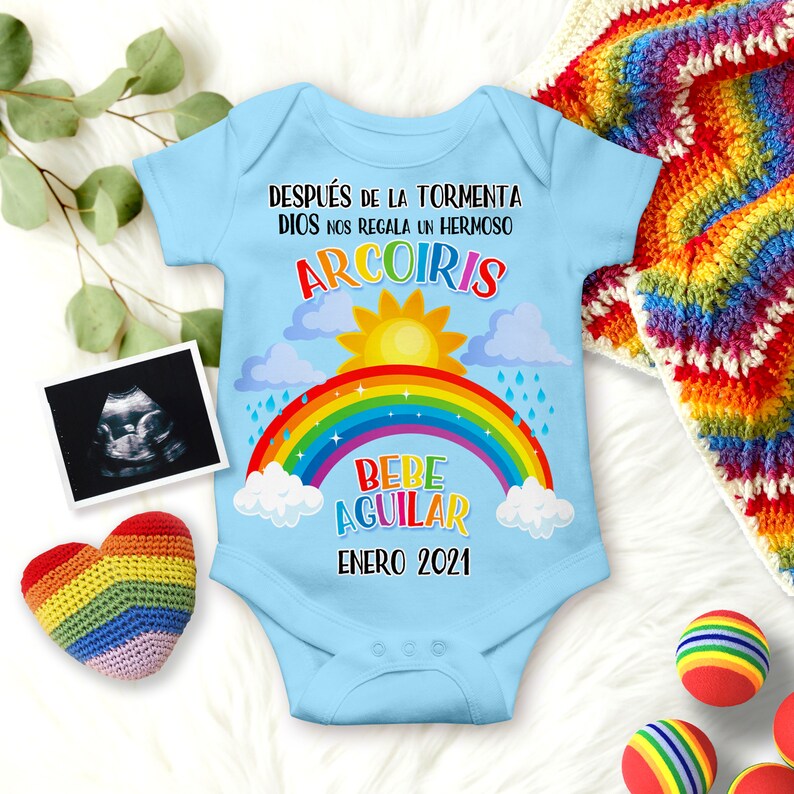 May include: A light blue baby onesie with a rainbow, clouds, and sun graphic. The text on the onesie reads "Despues de la tormenta Dios nos regala un hermoso arcoiris Bebe Aguilar Enero 2021".