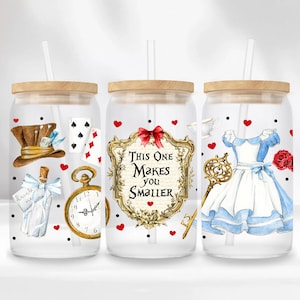 Puede incluir: Tres vasos de vidrio transparente con tapas de madera y pajitas. Cada vaso presenta un diseño diferente con temática de Alicia en el País de las Maravillas. El primer vaso tiene un sombrero de Sombrerero Loco, cartas de juego y un reloj de bolsillo. El segundo vaso tiene un marco dorado con el texto "This One Makes You Smaller". El tercer vaso tiene una llave, una taza de té y Alicia con su vestido azul.
