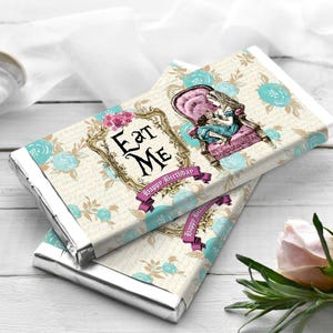 Puede incluir: Dos barras de chocolate con envoltorio de papel de aluminio plateado. Los envoltorios presentan un tema de Alicia en el país de las maravillas con las palabras "Eat Me" en un marco decorativo, un personaje sentado y "Happy Birthday" en una pancarta. Detalles florales y de escritura.
