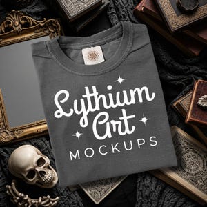Puede incluir: Una camiseta gris oscuro con el texto blanco "Lythium Art Mockups". La camiseta está rodeada de libros antiguos, una calavera y un espejo decorativo, creando una estética gótica o vintage.