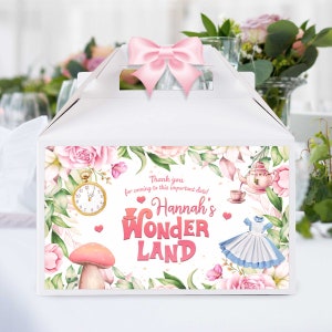 Puede incluir: Caja de regalo blanca con un lazo rosa. La caja está decorada con ilustraciones florales y el texto "Hannah's Wonderland". El diseño incluye un reloj de bolsillo, una seta y un vestido de Alicia en el país de las maravillas.