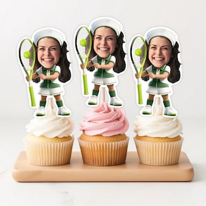 Puede incluir: Tres adornos para cupcakes con una imagen de dibujos animados de una persona sosteniendo una raqueta de tenis y una pelota. Los adornos están encima de cupcakes con glaseado blanco y rosa.