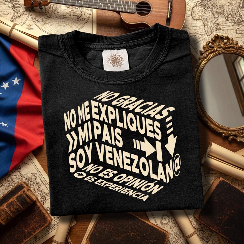 以下が含まれることがあります： 黒いTシャツ。白い3Dフォントで「NO GRACIAS, NO ME EXPLIQUES, MI PAIS, SOY VENEZOLANO, NO ES OPINION, ES EXPERIENCIA」と書かれています。ベネズエラの国旗とウクレレが置かれた木製の表面にTシャツがあります。