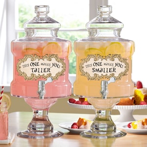 Puede incluir: Dos dispensadores de bebidas de vidrio transparente llenos de bebidas rosas y amarillas, cada uno con rodajas de limón. Los dispensadores tienen un grifo plateado y una etiqueta que dice "This one makes you taller" y "This one makes you smaller".