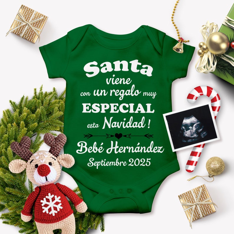 May include: Green baby onesie with white text that reads "Santa viene con un regalo muy ESPECIAL esta Navidad! Beb&eacute; Hern&aacute;ndez Septiembre 2025". The onesie is surrounded by Christmas decorations, including a candy cane, a reindeer toy, and a gift box.