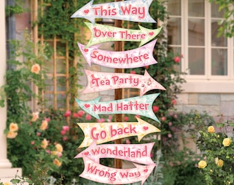 May include: A whimsical wooden signpost with colorful arrow signs pointing in different directions. Each sign features text like "This Way," "Over There," and "Wonderland" in a playful font, with floral and heart accents. The signpost is set in a garden.