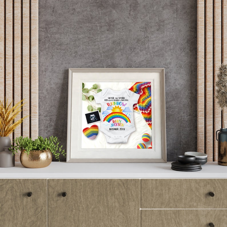 May include: A framed photo of a baby onesie with a rainbow design and the text "After the storm God always promises a beautiful rainbow" and "Baby Jackson December 2021". The onesie is surrounded by a rainbow crochet blanket, a heart-shaped ornament, and a sonogram photo.
