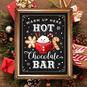 Puede incluir: Un letrero de pizarra enmarcado en oro con el texto "Warm Up Here Hot Chocolate Bar" en letras blancas. El letrero presenta una ilustración de dibujos animados de una taza de chocolate caliente con crema batida y una cara de muñeco de nieve.