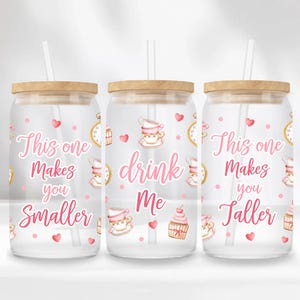 Puede incluir: Tres vasos de vidrio transparente con tapas y pajitas de bambú. Cada vaso tiene una cita diferente en escritura rosa: "This one Makes you Smaller", "drink Me" y "This one Makes you Taller". Los vasos están decorados con tazas de té, corazones y relojes.