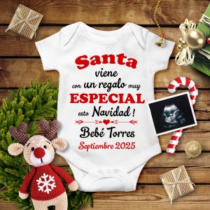 May include: White baby onesie with red text that reads "Santa viene con un regalo muy ESPECIAL esta Navidad! Bebe Torres Septiembre 2025". The onesie is surrounded by Christmas decorations, including a candy cane, a small Christmas tree, and a wrapped gift.