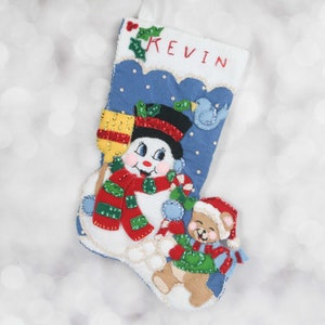 May include: A blue felt Christmas stocking with a snowman and a teddy bear holding a candy cane. The stocking has the name "Kevin" embroidered on it.
