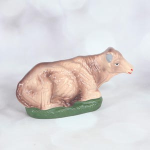 May include: A brown and white ceramic cow figurine with a green base. The cow is lying down with its head turned to the side. The cow has a pink nose and a blue ear.