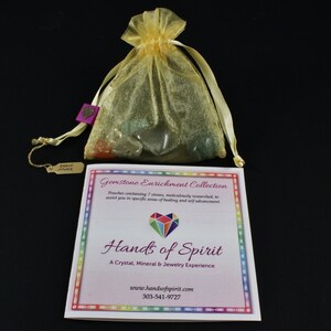 May include: A small yellow organza bag filled with colorful gemstones. The bag is tied with a ribbon and has a small tag attached to it. The tag says "Gemstone Enrichment Collection" and has a rainbow border. The text "Hands of Spirit" is printed in a large font below the tag. The text "A Crystal, Mineral & Jewelry Experience" is printed below the text "Hands of Spirit". The text "www.handsofspirit.com" and "303-541-9727" are printed below the text "A Crystal, Mineral & Jewelry Experience".