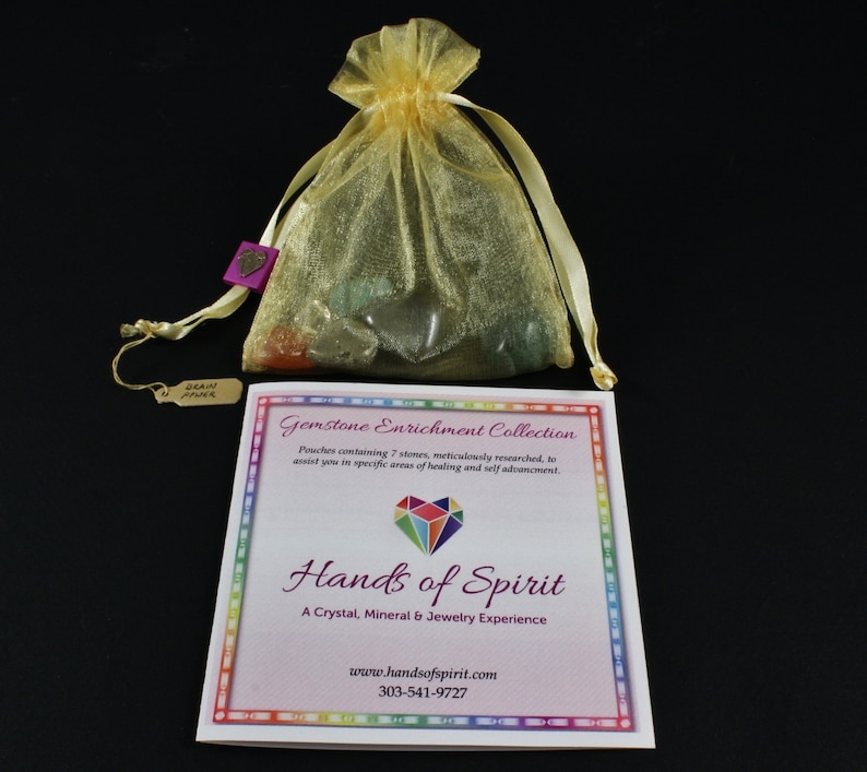 May include: A small yellow organza bag filled with colorful gemstones. The bag is tied with a ribbon and has a small tag attached to it. The tag says "Gemstone Enrichment Collection" and has a rainbow border. The text "Hands of Spirit" is printed in a large font below the tag. The text "A Crystal, Mineral & Jewelry Experience" is printed below the text "Hands of Spirit". The text "www.handsofspirit.com" and "303-541-9727" are printed below the text "A Crystal, Mineral & Jewelry Experience".