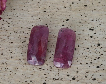May include: Two rectangular pink gemstones with a faceted cut. The gemstones are on a light brown wooden surface.