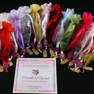 May include: A collection of 12 small, colorful organza bags tied with ribbons, each containing a crystal. The bags are arranged in a semi-circle on a black surface. A brochure with the text "Gemstone Enrichment Collection" and "Hands of Spirit" is in the center of the image.