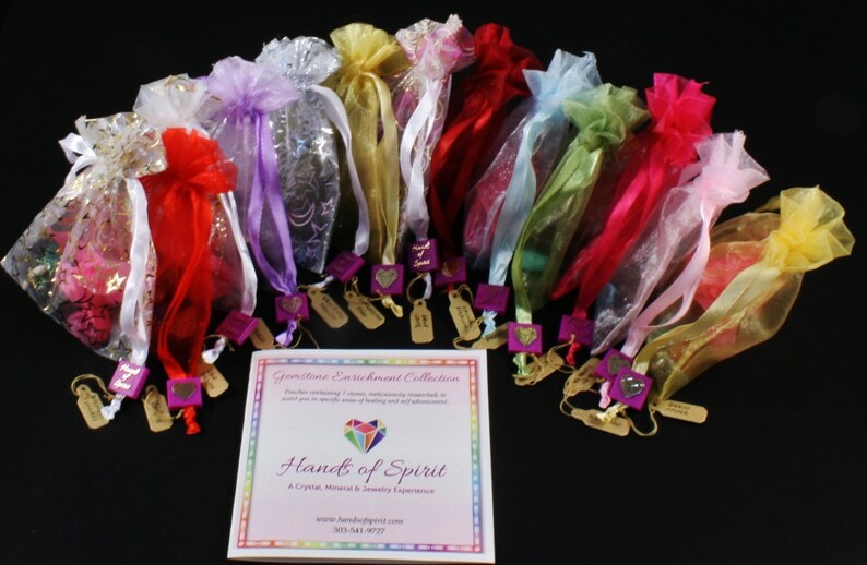 May include: A collection of 12 small, colorful organza bags tied with ribbons, each containing a crystal. The bags are arranged in a semi-circle on a black surface. A brochure with the text "Gemstone Enrichment Collection" and "Hands of Spirit" is in the center of the image.