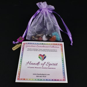 Puede incluir: Una bolsa de cordón morado llena de siete piedras preciosas de colores. La bolsa está etiquetada como "Gemstone Enrichment Collection" y es parte de la marca "Hands of Spirit". El texto en la tarjeta dice "Bolsas que contienen 7 piedras, meticulosamente investigadas, para ayudarte en áreas específicas de curación y auto-avance."