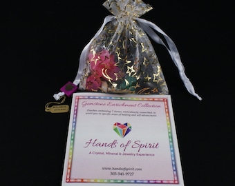 May include: A small white organza bag with gold stars and a pink heart charm. The bag contains a collection of gemstones. A card with the text "Gemstone Enrichment Collection" and "Hands of Spirit" is in front of the bag.