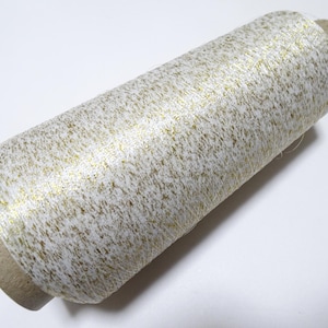 Puede incluir: Una bobina de hilo blanco con motas doradas. El hilo está enrollado alrededor de un tubo de cartón.