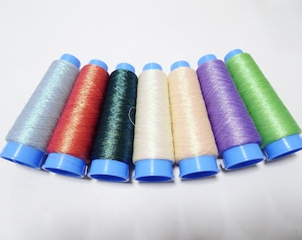May include: Seven spools of colorful thread in a row, each with a blue base. The thread colors include silver, red, dark green, cream, peach, purple, and green. These spools are likely used for sewing, embroidery, or other crafts.