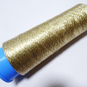 May include: A spool of metallic gold thread. The thread is tightly wound around a blue plastic core. The thread has a shiny, textured appearance, reflecting light. The spool is set against a white background.