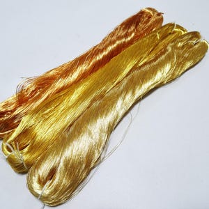 Puede incluir: Tres madejas de hilo metálico en tonos dorados y cobrizos. Los hilos están bien enrollados y tienen una superficie brillante y reflectante. Las madejas están dispuestas en diagonal sobre un fondo blanco, mostrando los diferentes colores y texturas.