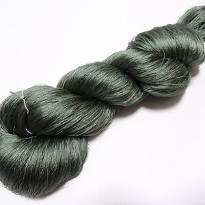 Pode incluir: Um novelo de fio verde escuro, bem enrolado e torcido, sobre um fundo branco. O fio parece ser feito de um material fino e sedoso, com um ligeiro brilho. A textura geral é suave e densa, sugerindo um material artesanal de alta qualidade.
