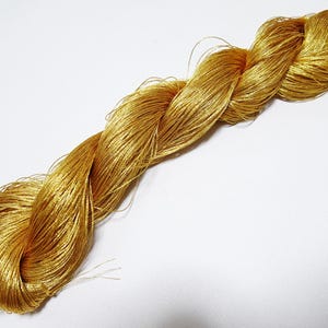 May include: A skein of golden-yellow embroidery floss, tightly twisted and lying diagonally across a white surface. The floss has a metallic sheen, suggesting a silk or rayon material, and is ideal for embroidery and other crafts.