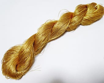 May include: A skein of golden-yellow embroidery floss, tightly twisted and lying diagonally across a white surface. The floss has a metallic sheen, suggesting a silk or rayon material, and is ideal for embroidery and other crafts.