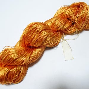 Puede incluir: Un ovillo de hilo naranja dorado, bien enrollado y texturizado, con una etiqueta en blanco adjunta. El hilo parece ser seda o una fibra fina similar, ideal para tejer u otras manualidades. El color es un tono rico y cálido.