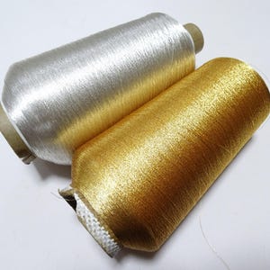 May include: Two spools of metallic thread, one silver and one gold, are displayed. The threads are tightly wound around cardboard cores. The silver thread has a bright, reflective sheen, while the gold thread has a warm, shimmering appearance. The threads are likely used for embroidery or decorative sewing.
