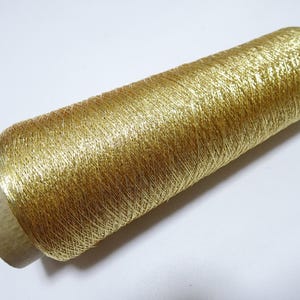 Pode incluir: Um carretel de fio dourado metálico. O fio é enrolado em torno de um núcleo de cartão, com uma tampa verde. O fio tem uma aparência brilhante e texturizada, adequado para projetos de bordado ou costura decorativa. O carretel tem cerca de 10 cm de comprimento.