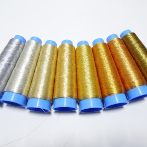 Könnte beinhalten: Acht Spulen mit Metallic-Garn in Silber- und Goldtönen. Die Spulen sind in einem Halbkreis angeordnet, wobei die silbernen Spulen links und die goldenen Spulen rechts liegen. Die Spulen haben blaue Plastikkappen.
