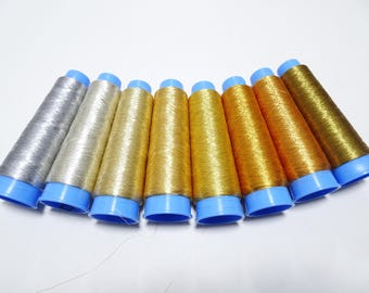 May include: Eight spools of metallic thread in shades of silver and gold. The spools are arranged in a semi-circle with the silver spools on the left and the gold spools on the right. The spools have blue plastic caps.