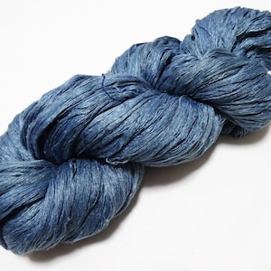 May include: A skein of blue yarn, tightly wound and textured. The yarn appears to be made of a natural fibre, with variations in the blue dye creating depth and visual interest. The yarn is on a white background.