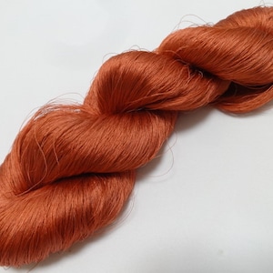 Puede incluir: Un ovillo de hilo de color óxido, hecho de una fibra natural suave. El hilo está retorcido y anudado, creando un aspecto texturizado.