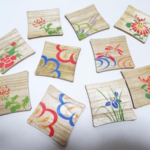 Può includere: Una collezione di dieci sottobicchieri quadrati in legno, ognuno caratterizzato da un design unico dipinto a mano. I motivi includono variopinti disegni floreali, forme astratte a onda e una libellula, dipinti su una venatura di legno chiaro.