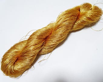 May include: A skein of golden-yellow yarn, tightly twisted and lying on a white surface. The yarn has a shimmering, metallic appearance, suggesting a silk or similar fiber. The strands are fine and delicate, with loose ends visible.