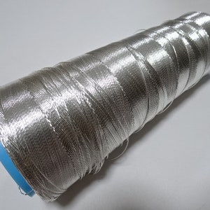 May include: A silver metallic thread spool. The thread is wound around a blue plastic core. The thread has a shiny, reflective appearance, possibly synthetic. The spool is on a plain white background. The thread is suitable for sewing and crafting.