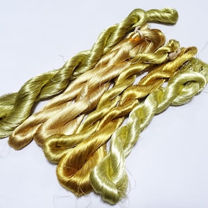 Puede incluir: Un conjunto de cinco madejas de hilo retorcido con un brillo lustroso en varios tonos de amarillo, dorado y verde oliva claro. Cada madeja está asegurada con un pequeño lazo, mostrando una textura suave y reflectante con un brillo vibrante.