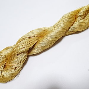May include: A skein of golden-yellow yarn, tightly twisted and coiled. The yarn appears to be made of fine, lustrous threads, reflecting light. The yarn is set against a plain white background, highlighting its texture and color. Ideal for crafting and textile projects.