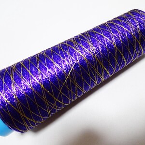 May include: A spool of metallic thread in a vibrant purple hue, interwoven with gold threads in a crisscross pattern. The thread is wound around a light blue plastic core. This decorative thread is suitable for various craft projects.